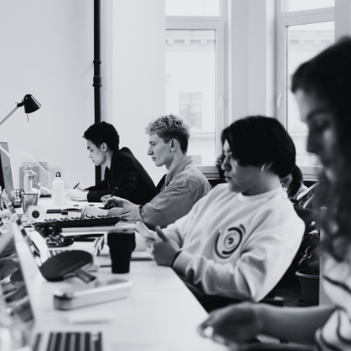 4 people lined up at individual desks, sitting and working on their computers. The image has a black and white filter on it.