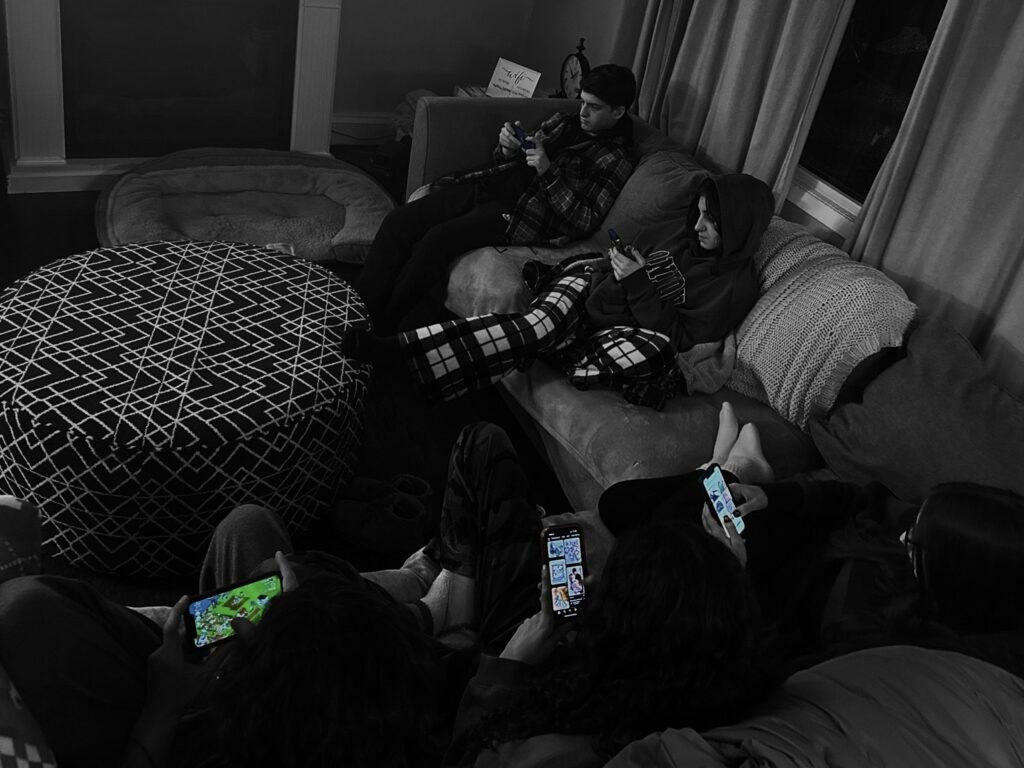 A group of friends are sitting on a couch. Each person is looking down at their cell phone. The image has a black and white filter on it and each phone is in color. 