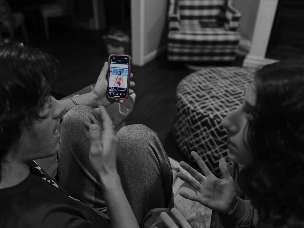 A young man and woman are sitting on a couch, seemingly arguing. He is holding his cell phone up, revealing an instagram post he has liked of a woman in a bathing suit. The image has a black and white filter on it and the cell phone is in color.