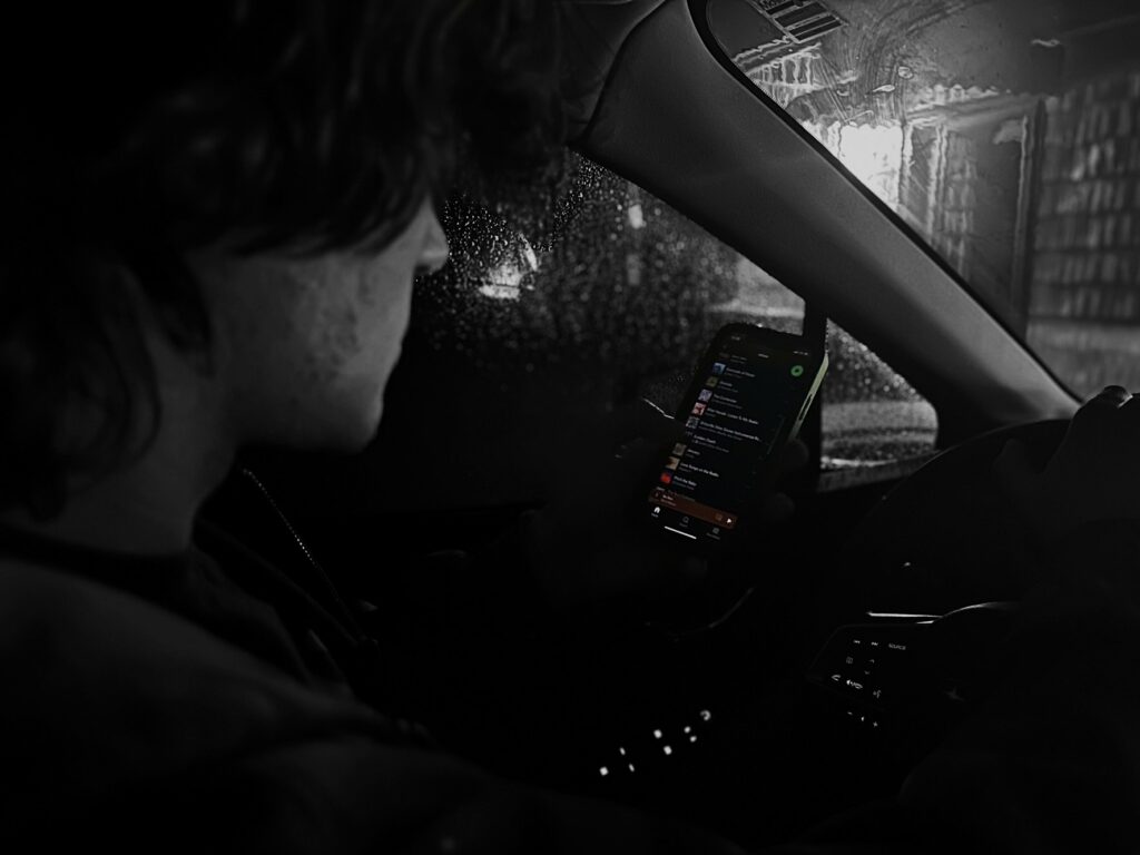 A man is in the driver's seat of a vehicle in motion. He is holding and scrolling though his cell phone while he drives. The image has a black and white filter on it and the cell phone is in color.