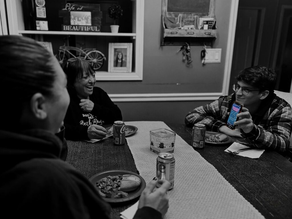 A teenage boy at the dinner table with his family, showing them a video on his cell phone. They are all laughing together. The image has a black and white filter on it and the cell phone is in color. 