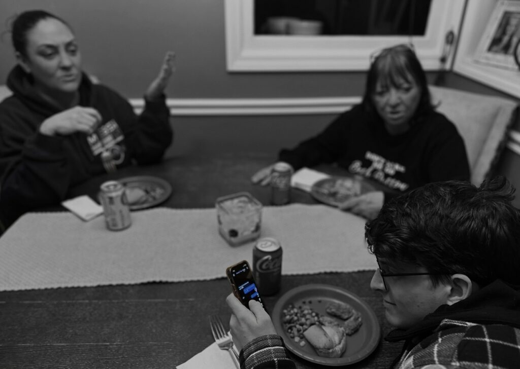 A teenage boy sitting at the dinner table with his family, scrolling on his cell phone while they are angrily trying to speak to him. The image has a black and white filter on it and the cell phone is in color.