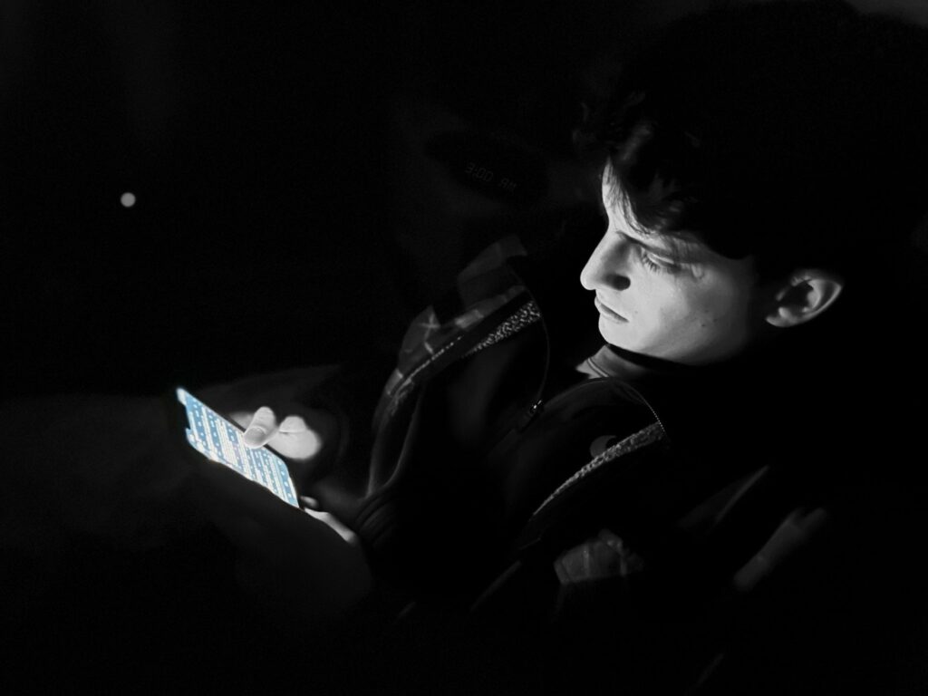 A young man is scrolling on his cell phone in bed. A clock in the background reads "3:00 AM". The image has a black and white filter on it, the cell phone is in color.