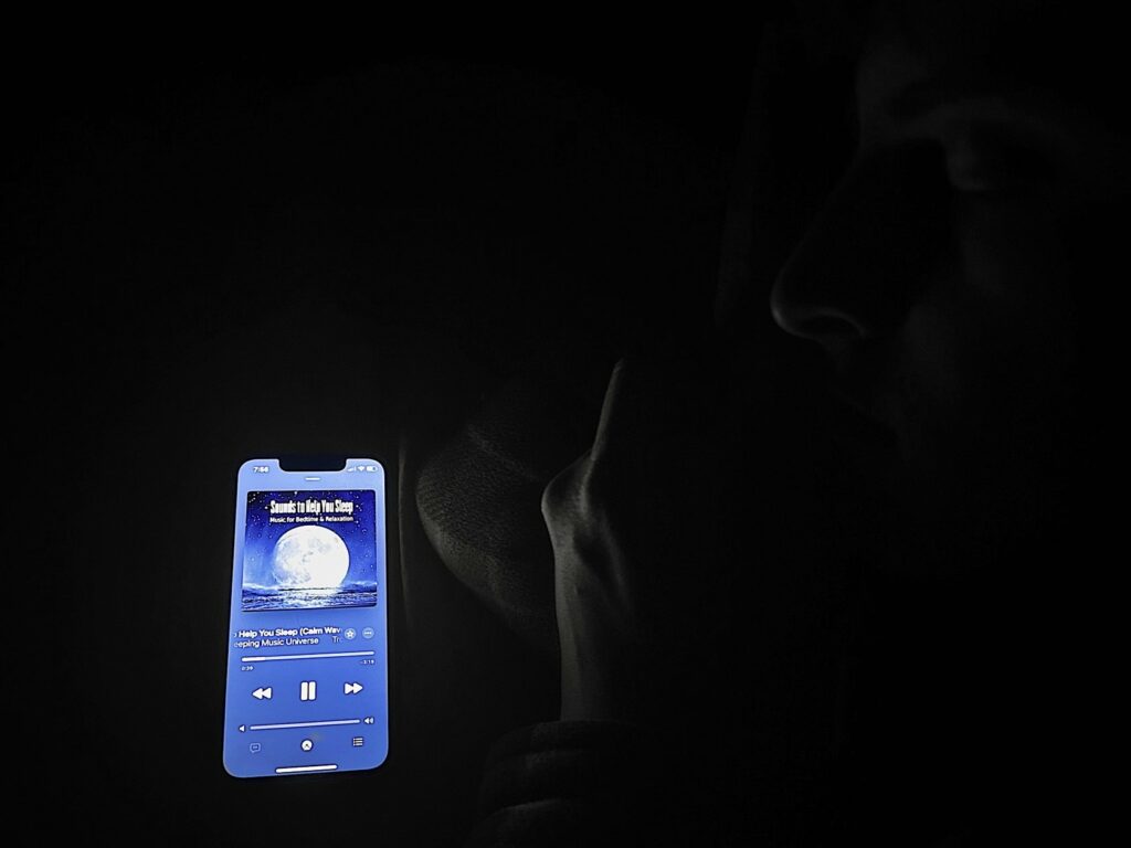 A young man is asleep next to his cell phone in bed. The cell phone is playing an album called "Sounds to Help You Sleep". The image has a black and white filter on it and the cell phone is in color.
