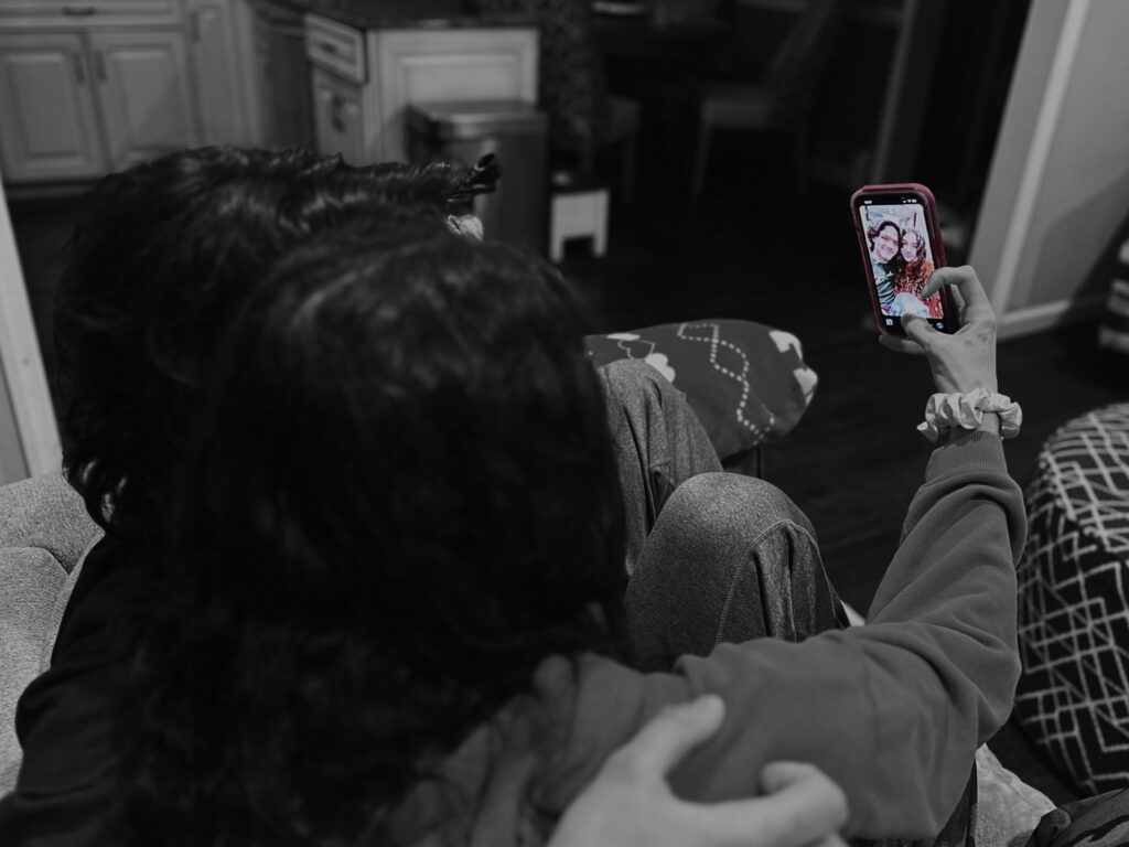A young man and woman sitting on the couch, cuddling and taking an Instagram photo on the woman's cell phone. The image has a black and white filter on it and the cell phone is in color.