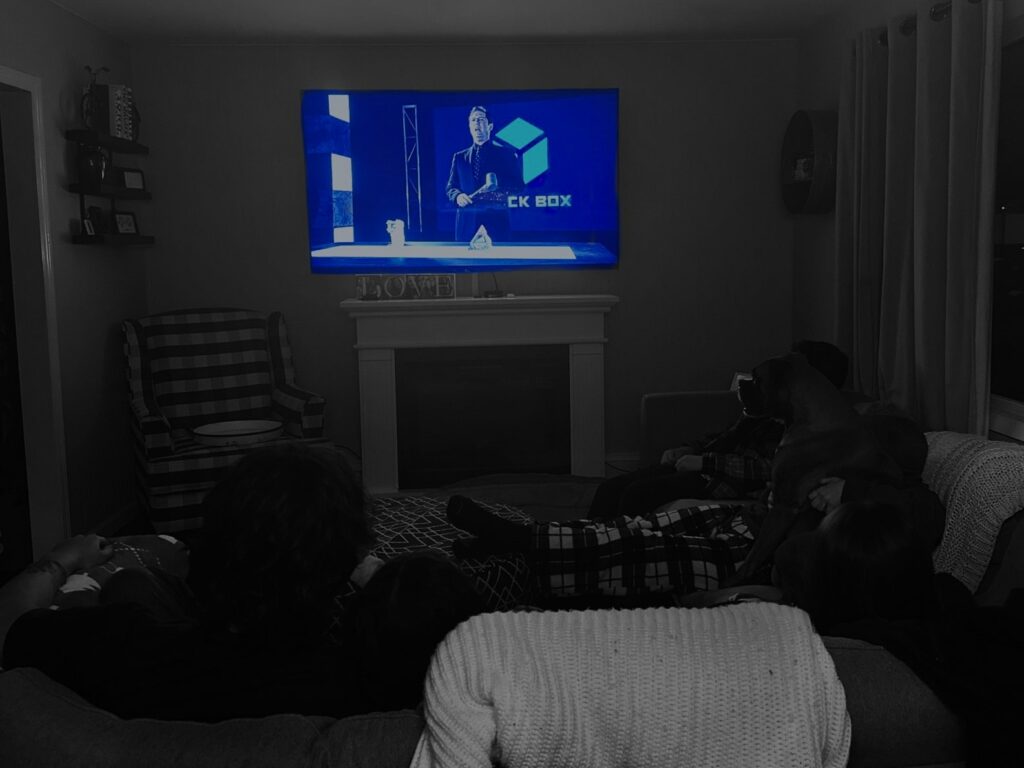 A group of friends are cuddling on the couch watching a movie on the television. The image has a black and white filter on it and the television is in color.