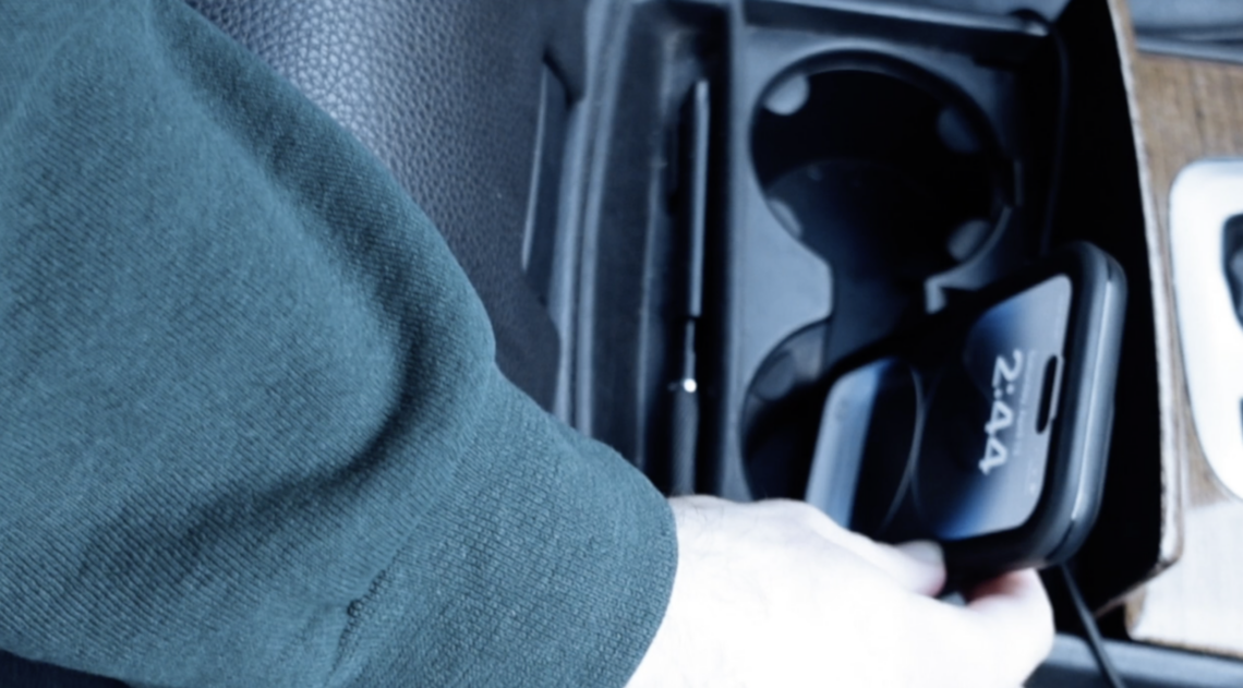 A man's arm reaching into the cup holder of a vehicle, holding a cell phone.